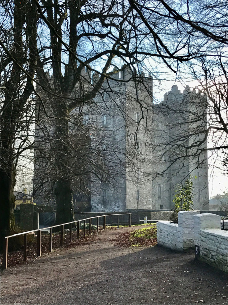 A grey stone castle is seen through the bare branches of trees.