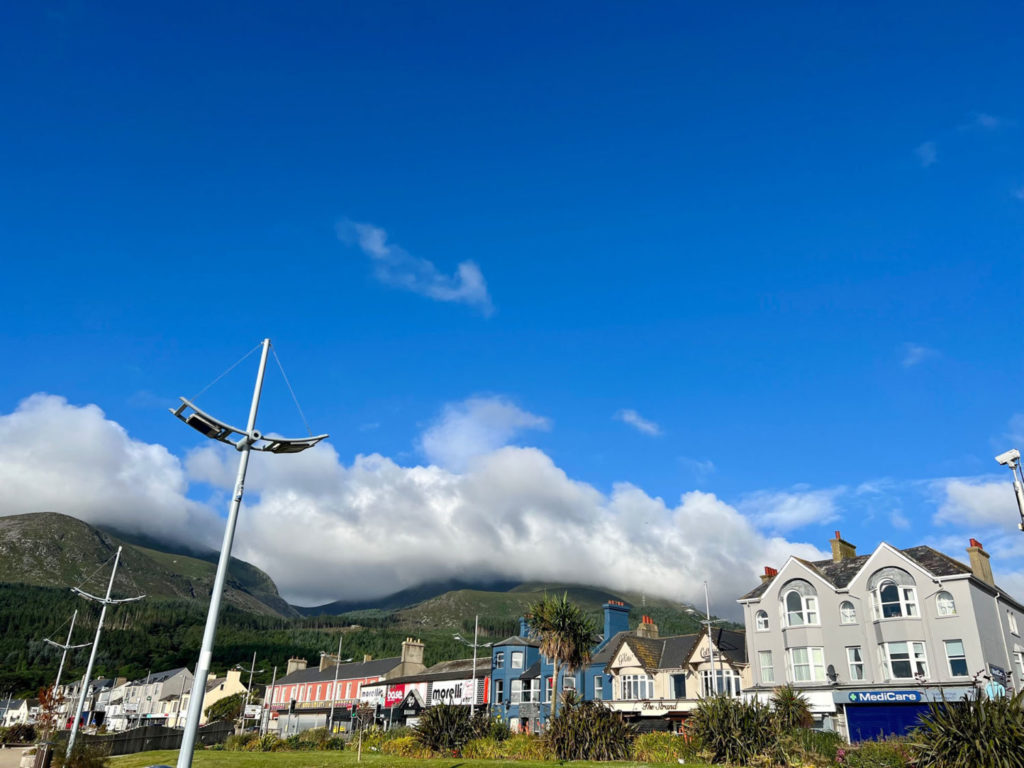 Clouds flow along the top of a mountain, with the seafront street of an Irish town in the foreground.