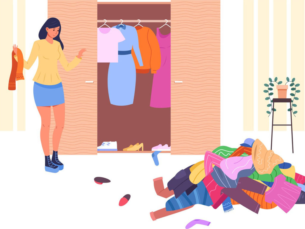 A woman stands next to an organized closet, near an untidy pile of clothes on the floor.