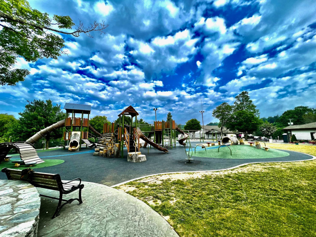 A variety of well-kept playground equipment on a rubbery surface in a park, with striking clouds in a vibrant blue sky.