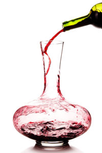 Red wine is being poured vigorously from a green bottle into a clear decanter. 