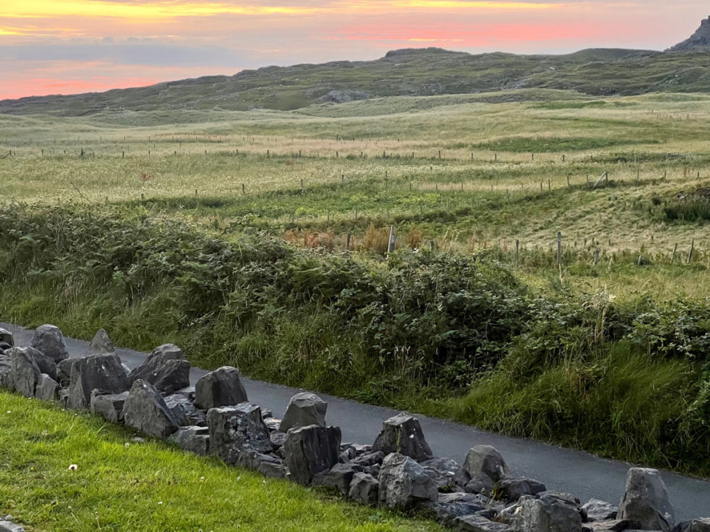 Sunset view of a field, across a narrow road, with the jagged top of a stone wall in the foreground.