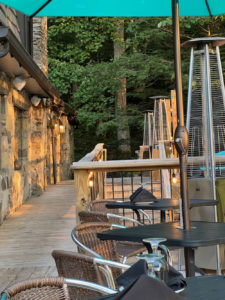 Tables and chair under an umbrella on a pleasant wooden deck outside of a stone building, with trees in the background.