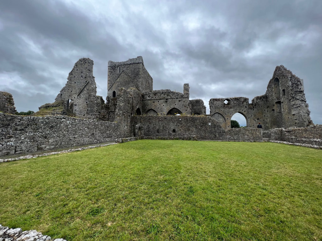 The ruins of a grey stone abbey, with a green field in the foreground and dark clouds overhead.