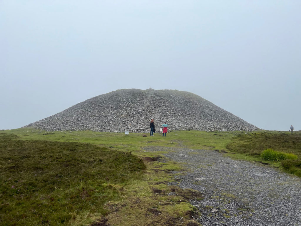 Maeve's Cairn is a megalithic tomb that consists of a large pile of rocks on top of a mountain. The two people standing in front of it look tiny by comparison.