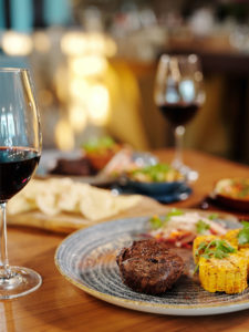 A glass of red wine sits on a table net to a plate containing a steak and grilled corn in the foreground; the background is artistically blurred but contains more food and wine.