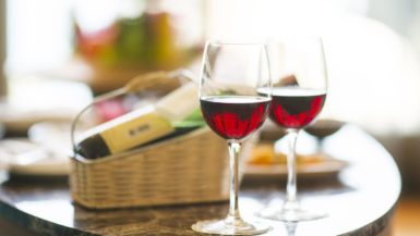 Wine glasses half-full of red wine in the foreground, with what appears to be a gift basket in the artistically blurred background