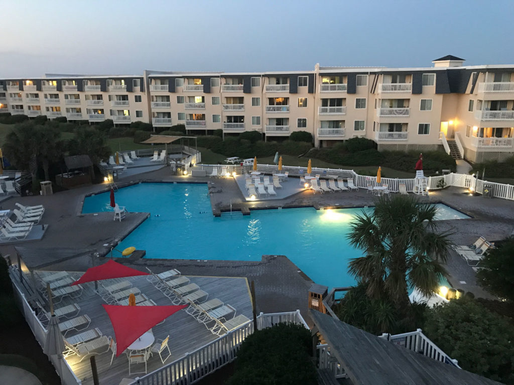 A beautiful outdoor swimming pool area, with lights, in the early evening, with a row of condos with balconies in the background, located in Atlantic Beach on the Crystal Coast.