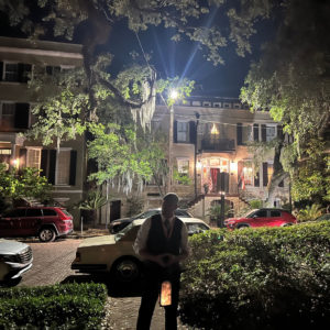 A man carrying a candle lantern is silhouetted by the lights from a historic mansion in Savannah