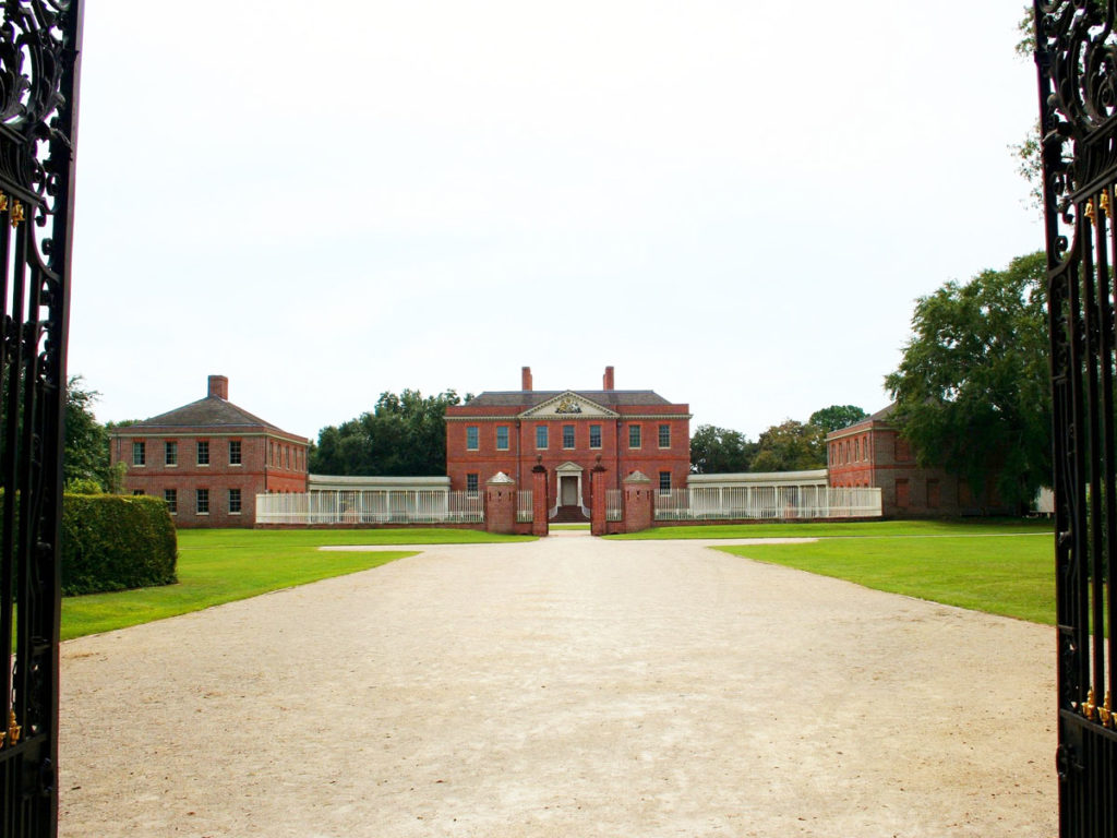 Tryon Palace, a large, Georgian-style red brick building, is seen through its gates. It is flanked by two simpler red brick outbuildings. It's located in New Bern, on the "Inner Banks" of the Crystal Coast.