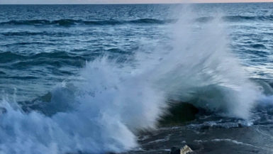 A wave crashes dramatically on a rock at the beach in front of a pastel sunset.