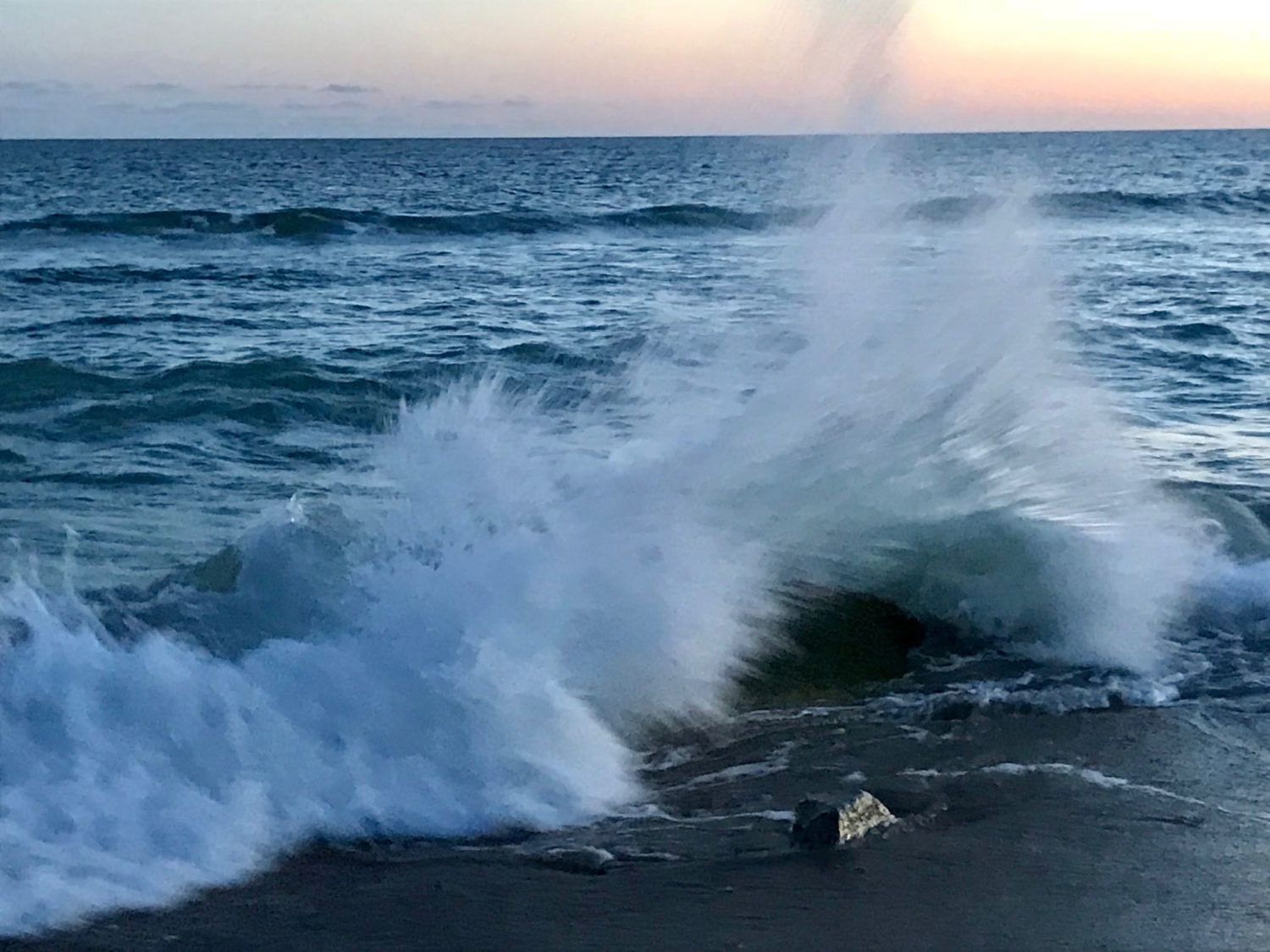 A wave crashes dramatically on a rock at the beach in front of a pastel sunset.