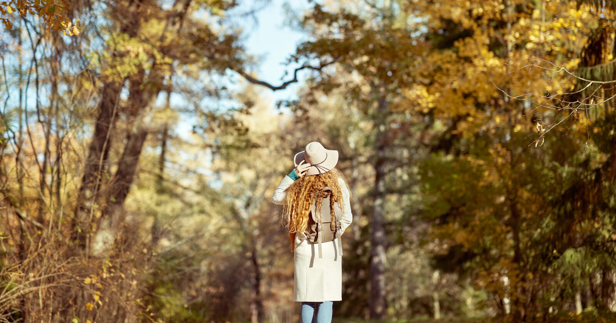 A red-haired woman in a stylish hat, raincoat, and backpack walks on a trail through trees in autumn.