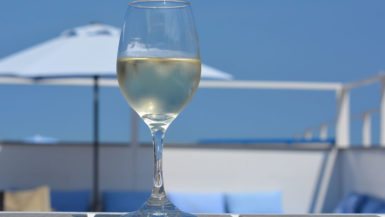 A cold glass of white Chardonnay wine stands on a picnic table in front of a beach umbrella and blue sky on a sunny day.