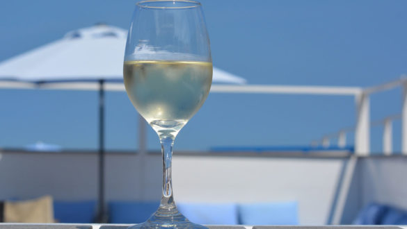 A cold glass of white Chardonnay wine stands on a picnic table in front of a beach umbrella and blue sky on a sunny day.