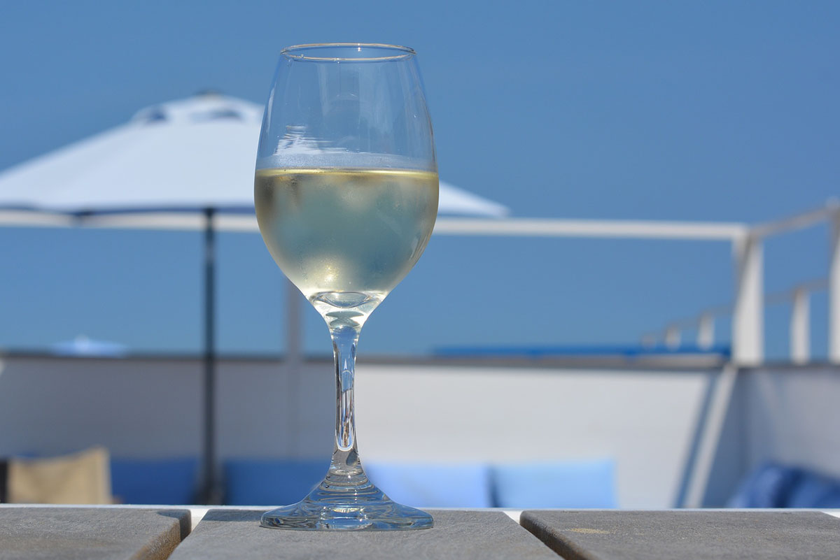 A cold glass of white Chardonnay wine stands on a picnic table in front of a beach umbrella and blue sky on a sunny day.