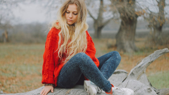 A blond woman in blue jeans, white sneakers, and a red sweater sits on a gray boulder amidst an artistically blurred cool-weather landscape.