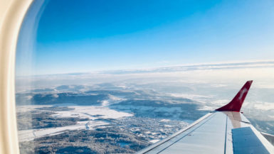 View out an airplane window, showing blue sky and a partly snowy landscape between the wing and the window frame.