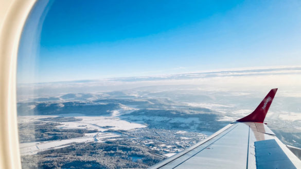 View out an airplane window, showing blue sky and a partly snowy landscape between the wing and the window frame.