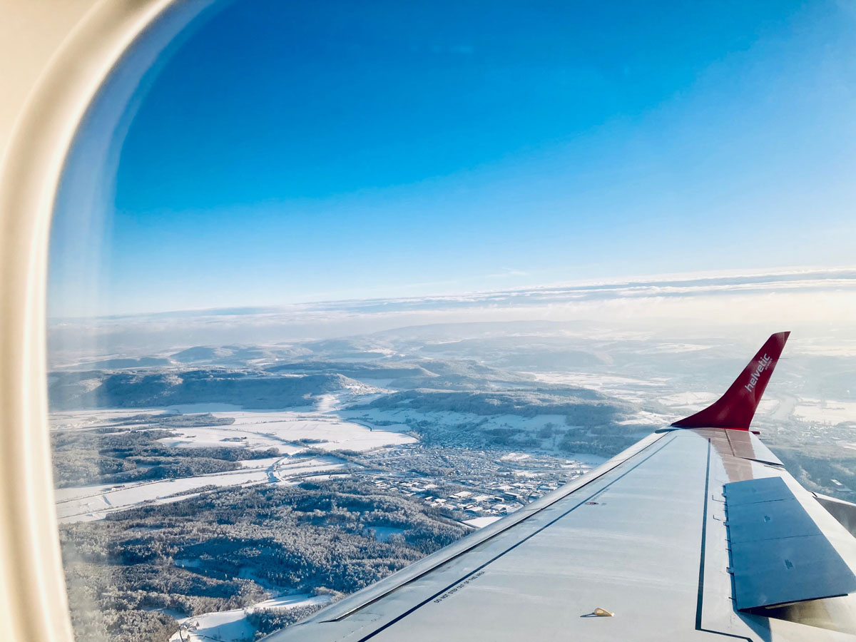 View out an airplane window, showing blue sky and a partly snowy landscape between the wing and the window frame.