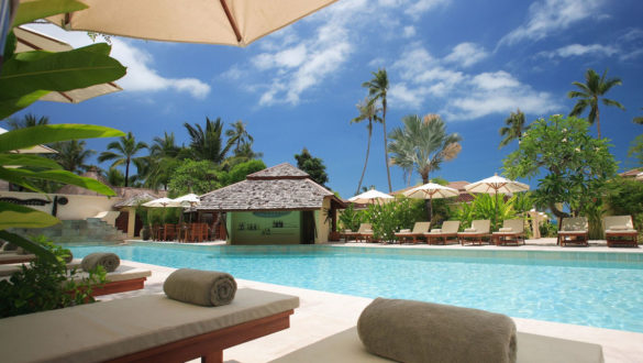 An umbrella shades comfortable lounges beside a swimming pool surrounded by palm trees beneath a sunny blue sky with a few puffy white clouds.