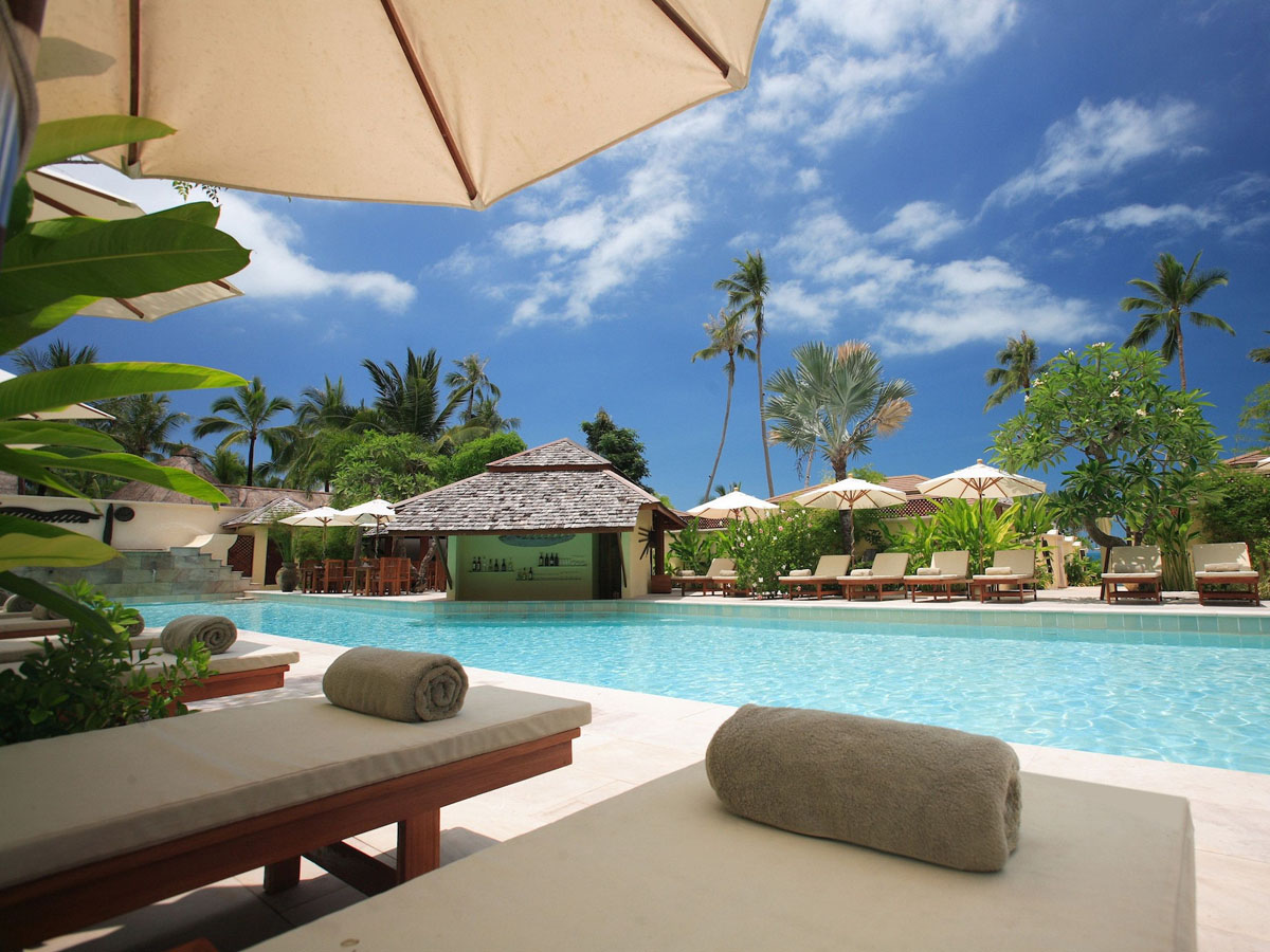 An umbrella shades comfortable lounges beside a swimming pool surrounded by palm trees beneath a sunny blue sky with a few puffy white clouds.